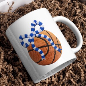 May include: White ceramic mug featuring a crocheted basketball design with black lines and an orange hue. A blue and white crocheted scarf is tied around the basketball. The mug is presented in a brown cardboard box.