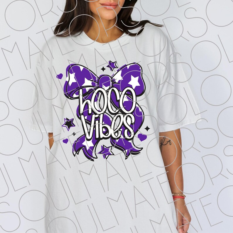 May include: White t-shirt featuring a purple bow design with white stars and the words "HOCO Vibes" in a stylized font. The design includes small star and heart accents. The shirt has short sleeves and a relaxed fit.