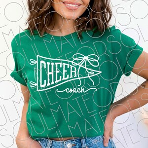 May include: A green t-shirt with rolled-up sleeves. The shirt features a white pennant design with the words "CHEER coach" in a stylized font. A bow and arrow design is above the word "CHEER".