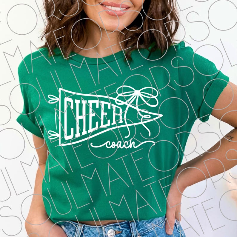 May include: A green t-shirt with rolled-up sleeves. The shirt features a white pennant design with the words "CHEER coach" in a stylized font. A bow and arrow design is above the word "CHEER".
