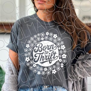 以下が含まれることがあります： 白い花で囲まれた、白い円のデザインに「Born to Thrift」という文字が入った濃い灰色のTシャツ。 シャツは袖をまくり、カジュアルで快適なスタイルです。