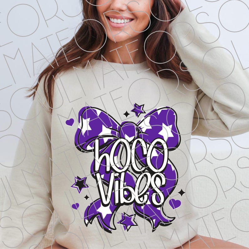 May include: A cream-colored sweatshirt featuring a large purple bow with white stars and the words "HOCO Vibes" in white. The design includes additional stars and hearts. The sweatshirt is a casual, comfortable style.
