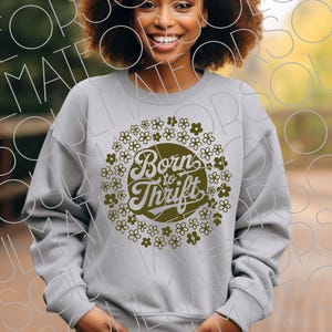 以下が含まれることがあります： 「Born to Thrift」の文字を囲むように花柄がデザインされた、ライトグレーのスウェットシャツ。 文字と花柄はくすんだオリーブグリーンです。 スウェットシャツはクルーネックです。