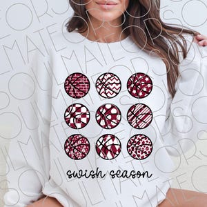 May include: White sweatshirt featuring nine basketball designs with various patterns in maroon and black. The phrase "swish season" is printed in a script font at the bottom. The sweatshirt is a casual, sporty style.