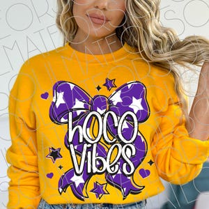 May include: Mustard yellow sweatshirt with a large purple bow design. The bow is decorated with white stars and the words "HOCO VIBES" in white and black. Small purple hearts and stars are scattered around the bow.