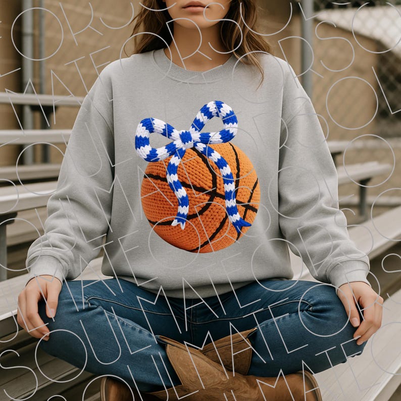 May include: A gray sweatshirt featuring a crocheted basketball with a blue and white striped bow. The basketball is orange with black lines. The sweatshirt is paired with blue jeans and brown boots.