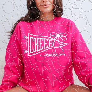 May include: A bright pink sweatshirt with the word "CHEER" in white script inside a pennant shape, with a bow and the word "coach" underneath. The sweatshirt is a solid color.
