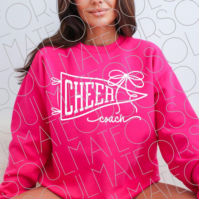 May include: A bright pink sweatshirt with the word "CHEER" in white script inside a pennant shape, with a bow and the word "coach" underneath. The sweatshirt is a solid color.