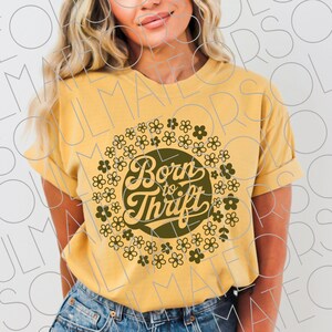 以下が含まれることがあります： マスタードイエローのTシャツ、袖は折り返されています。「Born to Thrift」の文字がレトロなフォントで書かれた円形のデザインで、小さな濃い緑色の花で囲まれています。柔らかい素材で作られたTシャツです。