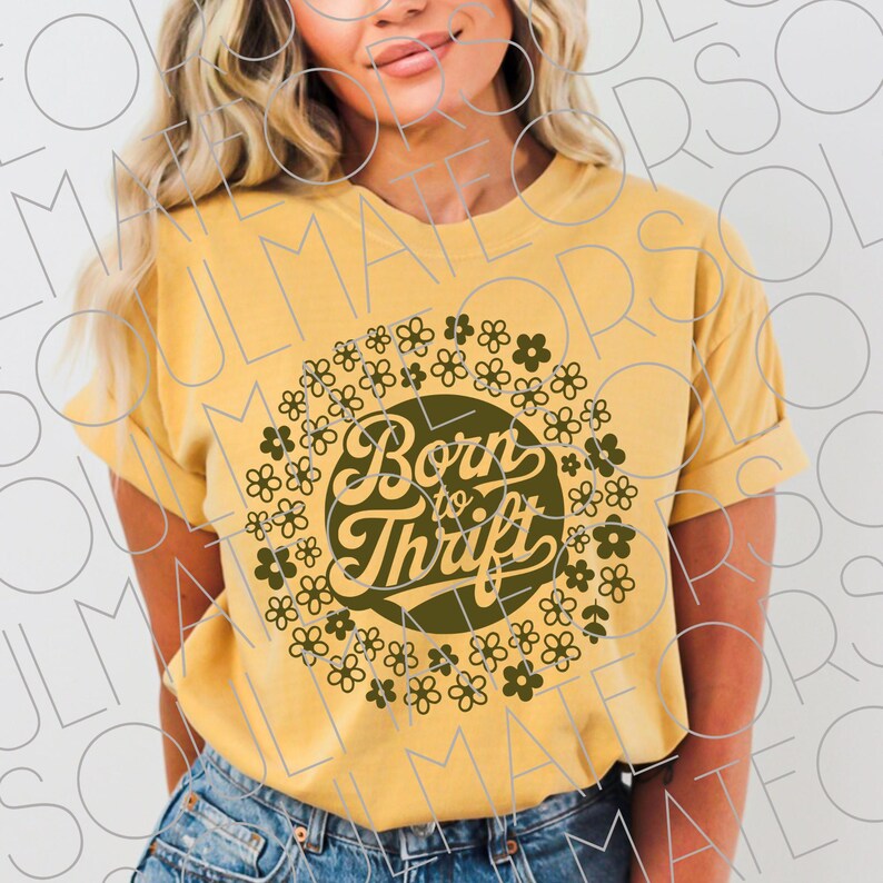 以下が含まれることがあります： マスタードイエローのTシャツ、袖は折り返されています。「Born to Thrift」の文字がレトロなフォントで書かれた円形のデザインで、小さな濃い緑色の花で囲まれています。柔らかい素材で作られたTシャツです。