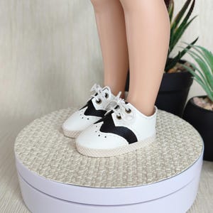 Könnte beinhalten: Weiße und schwarze Sattelschuhe mit weißen Schnürsenkeln. Die Schuhe haben einen schwarzen Akzent auf dem Spann und eine strukturierte, cremefarbene Sohle. Die Schuhe stehen auf einem weißen, drehbaren Präsentationsständer.