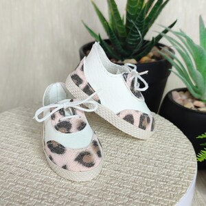 Könnte beinhalten: Ein Paar Miniatur-Sneaker in Weiß und Leopardenmuster mit weißen Schnürsenkeln. Die Schuhe haben eine weiße Zehenkappe und Ferse mit einem rosa-braunen Leopardenmuster auf Zehen und Seiten. Die Schuhe sind wahrscheinlich für eine Puppe.