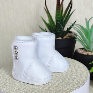 Puede incluir: Un par de botas blancas para bebé con forro de piel sintética. Las botas tienen tres botones plateados en el lateral y están hechas de un material liso similar al cuero. Las botas están diseñadas para bebés.