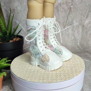 Könnte beinhalten: Weiße Schnürstiefel mit floralen Akzenten. Die Stiefel haben eine klobige Sohle und sind mit pastellfarbenen floralen Spitzeneinsätzen verziert. Die Stiefel werden auf einer weißen Plattform präsentiert, um ihr Design hervorzuheben.