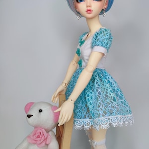 Puede incluir: Una muñeca con cabello azul y morado, vestida con un vestido de encaje azul y medias blancas. Un oso de peluche blanco con detalles en rosa está sentado en una silla de madera junto a la muñeca. La muñeca tiene ojos azules y lleva pendientes.
