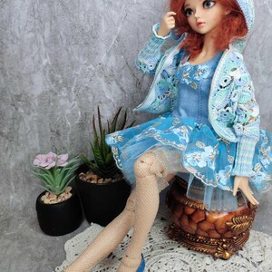 May include: A doll wearing a blue dress with floral embroidery, a matching crocheted jacket and hood, and blue shoes. The doll has red hair and is sitting on a decorative wooden stool. The doll is wearing fishnet stockings.