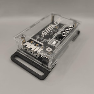 Könnte beinhalten: Ein transparentes Kunststoffgehäuse mit einer schwarzen Platine im Inneren. Die Platine trägt den Aufdruck "SIMAGIC". Das Gehäuse hat einen schwarzen Gurt.