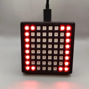 Könnte beinhalten: Ein schwarzes quadratisches Gerät mit einem Raster aus roten LED-Leuchten. Das Gerät ist mit einem schwarzen USB-Kabel verbunden.