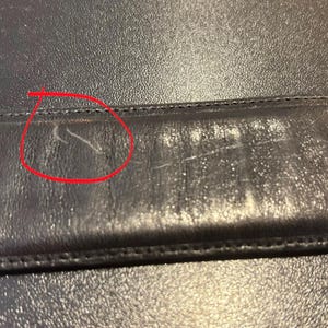 May include: Close-up of a black leather surface with visible scratches and wear. The leather appears to be slightly textured, with a stitched seam running horizontally across the image. The wear and tear suggest the item is used.