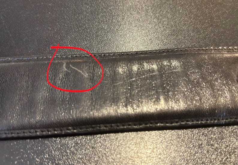 May include: Close-up of a black leather surface with visible scratches and wear. The leather appears to be slightly textured, with a stitched seam running horizontally across the image. The wear and tear suggest the item is used.