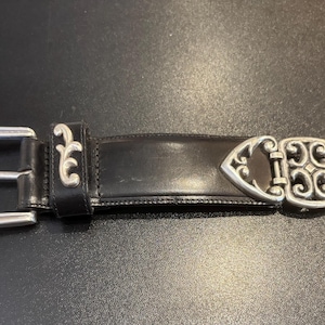 May include: Black leather belt with silver-colored metal buckle and decorative accents. The buckle is a classic design with a rectangular frame and a central bar. The belt has a decorative silver-colored metal clasp.