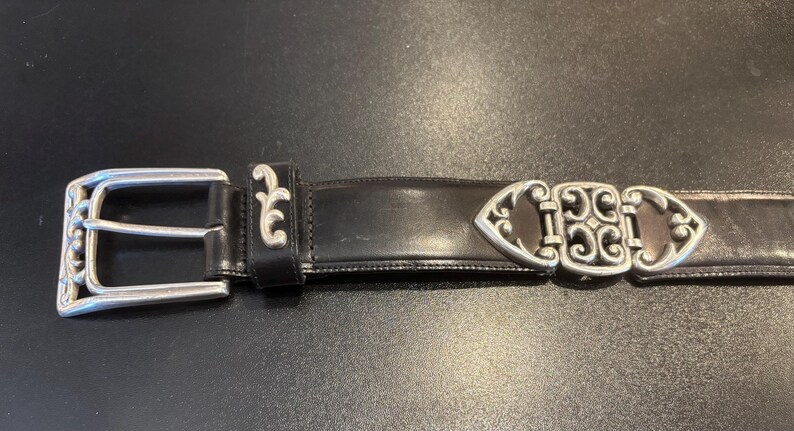 May include: Black leather belt with silver-colored metal buckle and decorative accents. The buckle is a classic design with a rectangular frame and a central bar. The belt has a decorative silver-colored metal clasp.