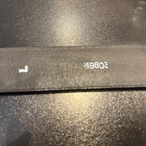 May include: Close-up of a black leather belt. The letter "L" and the number "49803" are printed in white on the belt. The belt is on a dark, textured surface.