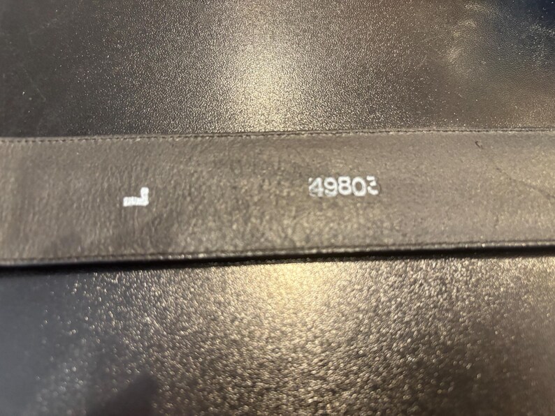 May include: Close-up of a black leather belt. The letter "L" and the number "49803" are printed in white on the belt. The belt is on a dark, textured surface.