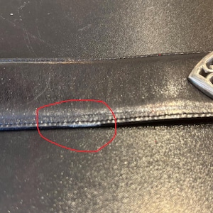 May include: Close-up of a black leather handbag. The leather has a textured appearance with visible stitching along the seams. A decorative silver-colored metal clasp is visible on the right side of the image.