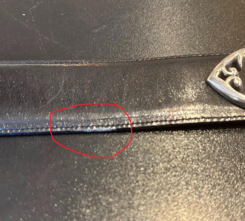 May include: Close-up of a black leather handbag. The leather has a textured appearance with visible stitching along the seams. A decorative silver-colored metal clasp is visible on the right side of the image.