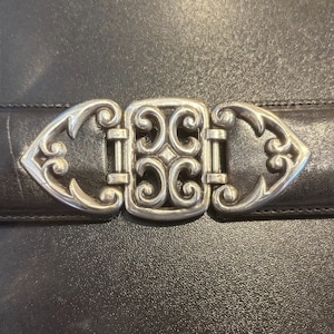 May include: Close-up of a black leather belt with a decorative silver-colored buckle. The buckle features an ornate, symmetrical design with scrolling details. The belt is likely for fashion or accessories.
