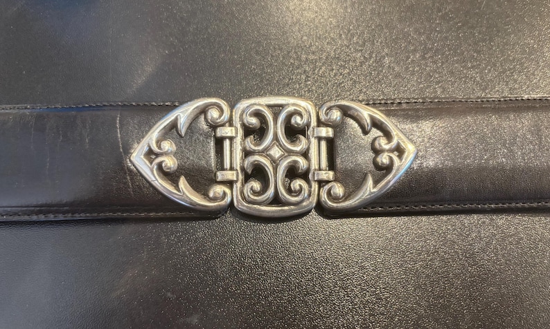 May include: Close-up of a black leather belt with a decorative silver-colored buckle. The buckle features an ornate, symmetrical design with scrolling details. The belt is likely for fashion or accessories.