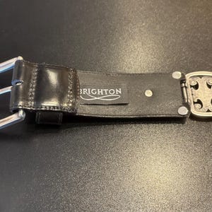 May include: Black leather belt with a silver-tone buckle and decorative clasp. The belt features the brand name "BRIGHTON" on a small tag. The buckle has a unique design.