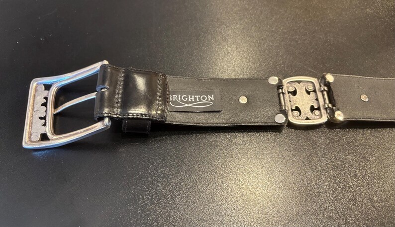 May include: Black leather belt with a silver-tone buckle and decorative clasp. The belt features the brand name "BRIGHTON" on a small tag. The buckle has a unique design.