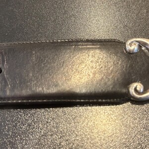 May include: A black leather belt with a decorative silver-colored buckle. The buckle has an ornate, curved design. The belt has several holes for adjustment.