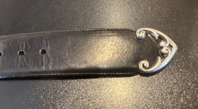 May include: A black leather belt with a decorative silver-colored buckle. The buckle has an ornate, curved design. The belt has several holes for adjustment.