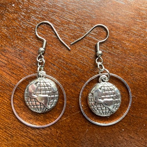 May include: A pair of silver-toned dangle earrings. Each earring features a globe charm suspended within a clear circular frame. The globe charm has detailed continents. The earrings have a hook closure.
