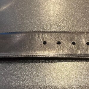 May include: Black leather belt with a decorative silver-colored buckle. The buckle has an ornate, swirling design. The belt has several holes for adjustment and shows signs of wear.