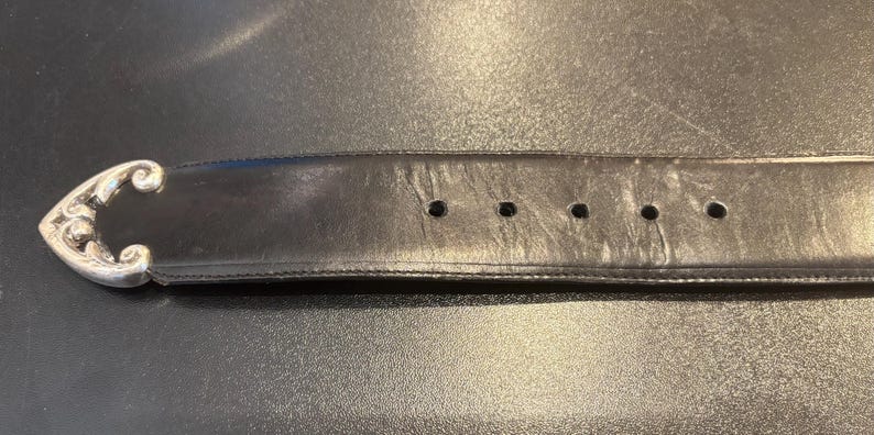 May include: Black leather belt with a decorative silver-colored buckle. The buckle has an ornate, swirling design. The belt has several holes for adjustment and shows signs of wear.