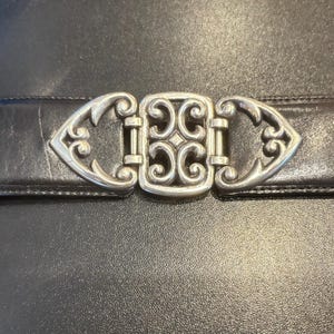 May include: A black leather belt with a decorative silver-colored buckle. The buckle has an ornate design with a central square and stylized end pieces. The belt is shown against a dark background.