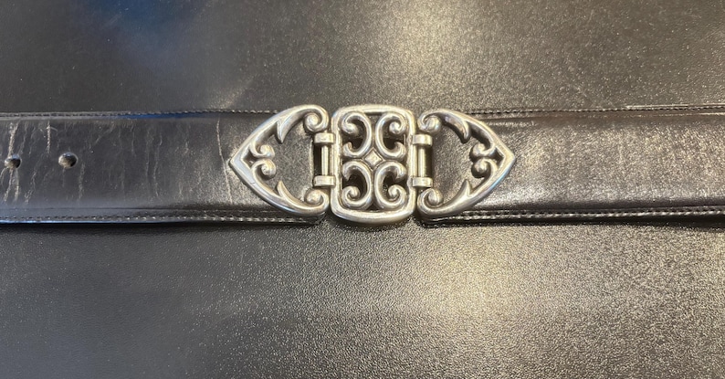 May include: A black leather belt with a decorative silver-colored buckle. The buckle has an ornate design with a central square and stylized end pieces. The belt is shown against a dark background.