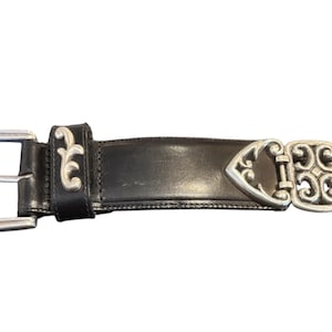 May include: Black leather belt with silver-tone buckle and decorative accents. The buckle is rectangular with a floral design. The belt has a silver-tone clasp with an ornate design.