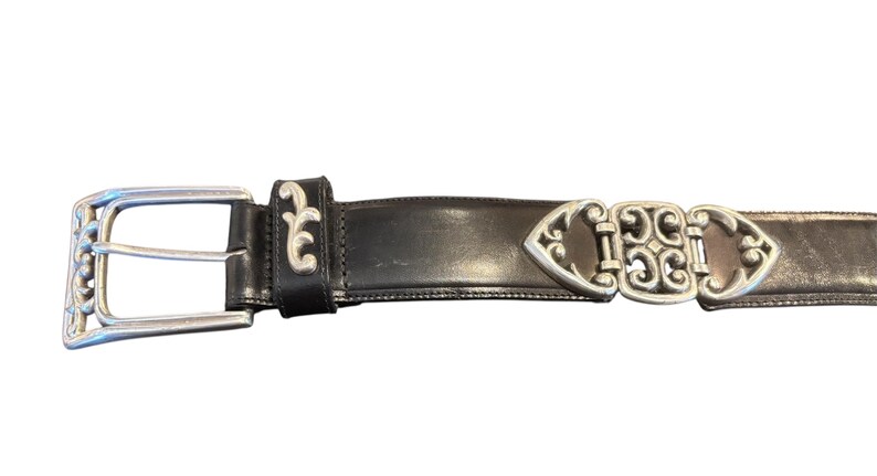 May include: Black leather belt with silver-tone buckle and decorative accents. The buckle is rectangular with a floral design. The belt has a silver-tone clasp with an ornate design.