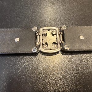 May include: A black leather belt with a silver-colored buckle. The buckle features a decorative cross design and is connected to the belt with small, silver-colored rivets. The belt is shown against a dark background.