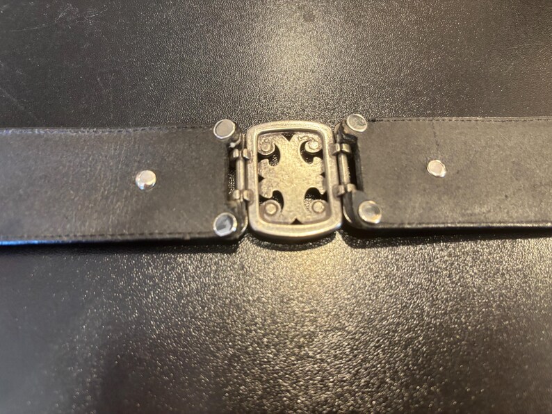 May include: A black leather belt with a silver-colored buckle. The buckle features a decorative cross design and is connected to the belt with small, silver-colored rivets. The belt is shown against a dark background.