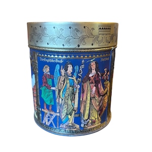 May include: A cylindrical metal tin featuring a gold-coloured lid with a leaf pattern. The blue body displays a colourful illustration of a historical scene with figures, including an angel, and the German text "Der Englische Gruß" and "Veit Stoß".