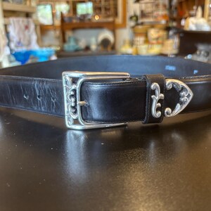 May include: Black leather belt with silver-toned decorative accents. The belt features a large, ornate buckle and decorative silver pieces along the strap. The belt is displayed on a dark surface.
