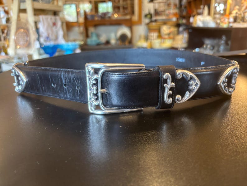 May include: Black leather belt with silver-toned decorative accents. The belt features a large, ornate buckle and decorative silver pieces along the strap. The belt is displayed on a dark surface.