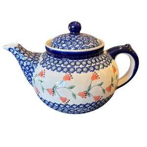 Op de afbeelding: Een keramische theepot met een crèmekleurig lichaam versierd met een patroon van oranje bloemen op groene ranken. Het deksel, de tuit en het handvat zijn voorzien van een donkerblauwe en lichtgroene stervormige geometrische rand.