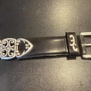 May include: Black leather belt with a silver-colored buckle and decorative accents. The belt features a detailed buckle design and a classic, elegant style. The belt is approximately 1.5 inches wide.